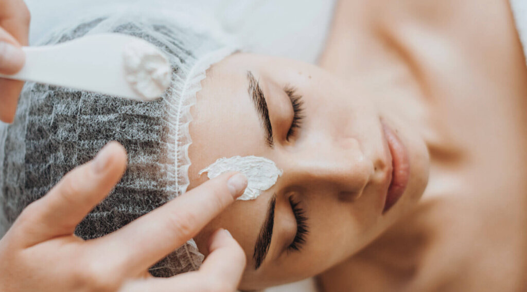 Cosmetician's hands applying facial cream mask on woman's forehead. Skin treatment. Spa beauty procedure